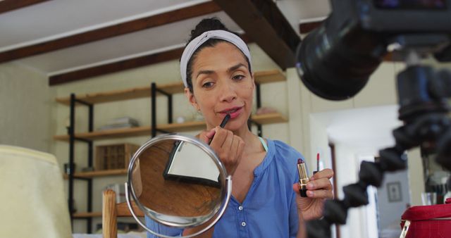 Beauty vlogger applying lipstick while being filmed by a camera in a home environment. Suitable for use in blogs, internet platforms, makeup tutorials, social media content, and advertisements focused on beauty, cosmetics, influencer marketing, and personal care.