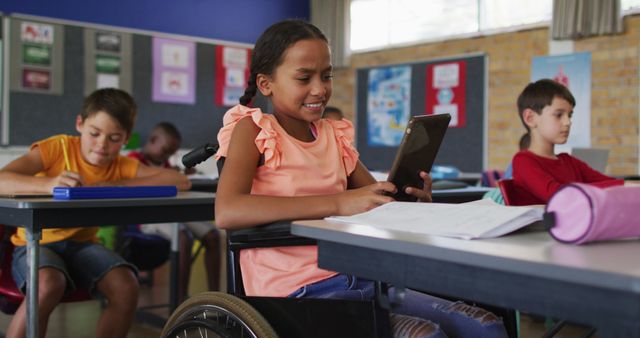 Image features a group of elementary school students engaging with technology and studying at their desks. A girl in a wheelchair is using a tablet, smiling. Other children are seated nearby, absorbed in their own work. Bright classroom with colorful educational posters on the wall. Ideal for concepts like inclusive education, modern learning environments, diversity in schools, and the use of technology in education.