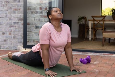 African american woman practicing cobra pose on brick patio with dumbbells, smartwatch