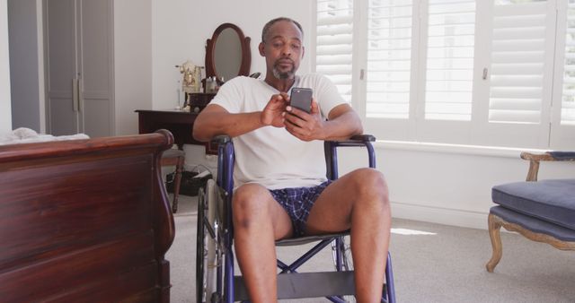 Thoughtful senior african american man sitting in wheelchair at home using smartphone. Communication, senior lifestyle, disability and domestic life.