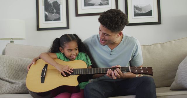 Father teaching daughter guitar on a cozy living room couch. Great for promoting family bonding, music education, and home activities. Perfect for blogs, ads, and educational content.