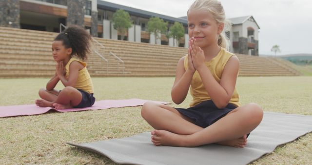 Image of focused diverse girls practicing yoga on mats in front of school. primary school education, sport and exercising concept.