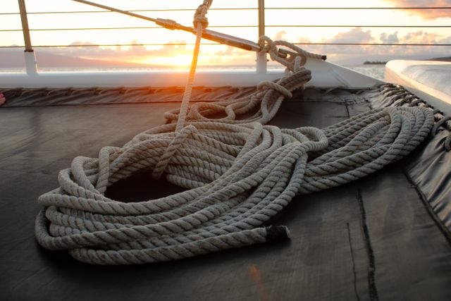 Coiled rope rests neatly on the deck of a yacht during sunset. The calm ocean and orange hues of the setting sun provide a peaceful vibe. Perfect for themes related to boating, nautical serenity, travel, and adventure.