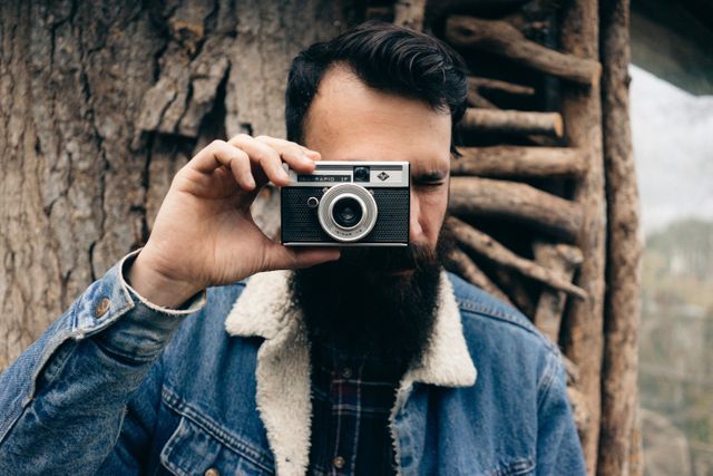 Man with a beard holding a vintage camera in front of his face, focusing outdoors against a background with a tree and rustic wooden structure. Suitable for use in projects related to photography, adventure, outdoor activities, and retro themes.