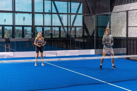 Mixed doubles padel practice on blue indoor court with glass walls and training partners