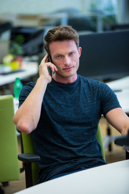 Young businessman in casual attire sitting at desk in modern office, using mobile phone for communication. Ideal for concepts related to business communication, professional environment, modern workplaces, and corporate lifestyle.