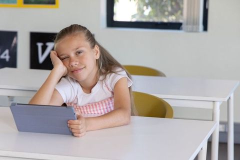 Preteen girl sitting in classroom using tablet and smiling, natural light digital learning