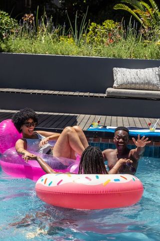 Friends laughing and floating on pink donut and translucent lounger in sunlit backyard pool