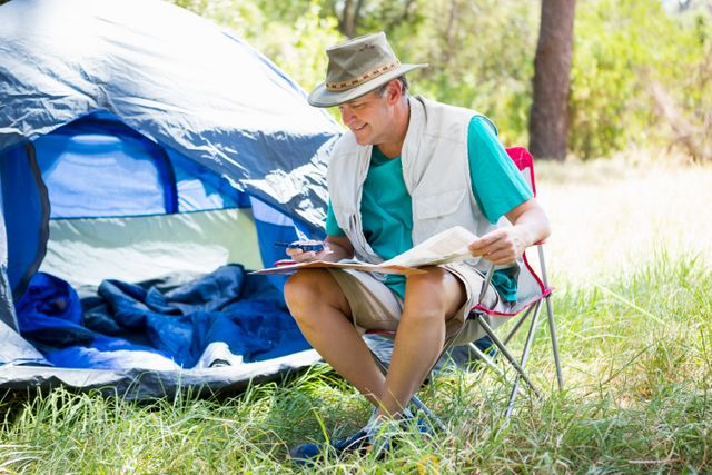 Senior man sitting on a chair beside his tent in a forest, reading a book and enjoying the outdoors. Ideal for use in articles or advertisements related to outdoor activities, camping, senior lifestyle, adventure travel, and nature exploration.
