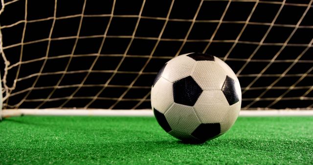 This image features a classic black and white soccer ball positioned on green turf with a goal net in the background, captured during nighttime. Ideal for use in sports magazines, promotional materials for football events, soccer training programs, and advertisements for sports equipment.