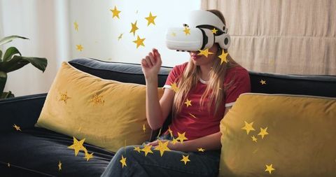 Young woman wearing white VR headset on sofa interacting with floating gold stars at home