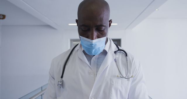 African American doctor wearing a protective face mask, looking down, with a stethoscope around his neck, in a well-lit hospital. Ideal for use in medical articles, healthcare promotions, hospital website content, health and safety guidelines, and COVID-19 awareness campaigns.
