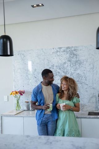 Diverse couple leaning and holding mugs at marble kitchen countertop, enjoying coffee