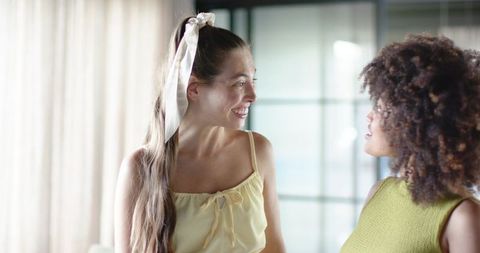 Smiling friends chatting in sunlit home interior wearing summer camisole and knit top
