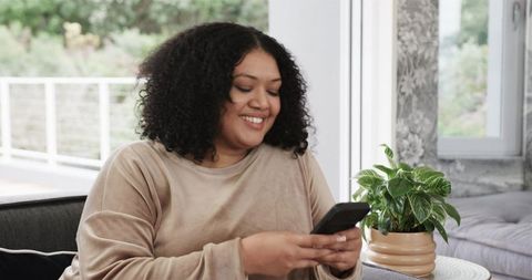 African american woman smiling and using smartphone on sofa in cozy modern living room