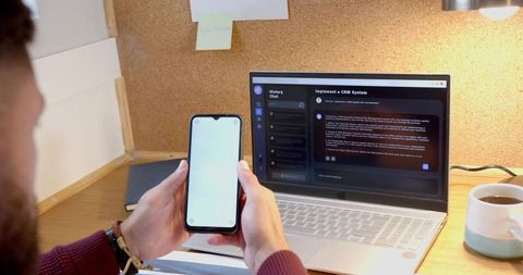 Bearded man holding smartphone while using laptop on wooden desk with corkboard and coffee