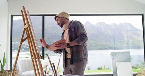 Young indian artist painting at easel with mountain and lake view in bright studio