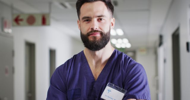 Male healthcare professional standing confidently in a hospital corridor, wearing purple scrubs and an ID badge. Suitable for use in healthcare, medical publications, hospital websites, health service promotions, and educational materials about healthcare professionals.