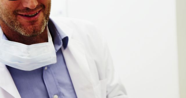 Shows close-up of a smiling doctor or healthcare professional wearing a white lab coat and a surgical mask in a medical setting. Ideal for medical blogs, healthcare promotional materials, and hospital websites to represent healthcare, professionalism, and friendliness.