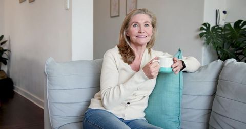 Senior Woman Relaxing on Sofa with Tea and Pillow, Showcasing a Cozy Home Setting