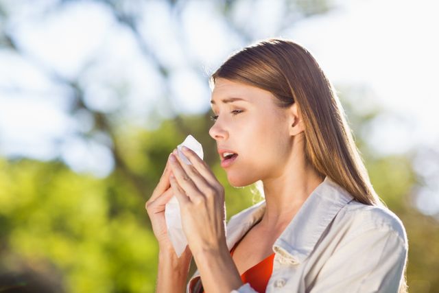 Young woman sneezing into a tissue while standing in a park. The background features green trees and sunlight, indicating a pleasant day. This image can be used for topics related to allergies, seasonal illnesses, health, and outdoor activities.