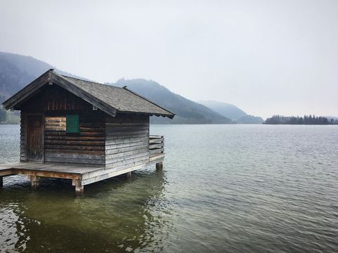 Weathered wooden boathouse sitting on stilts over calm lake with misty forested mountains