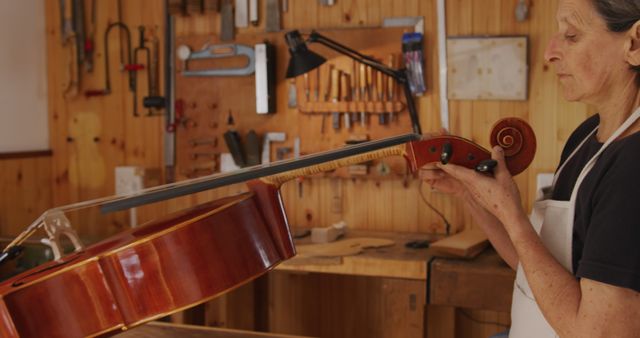 Side view of a senior Caucasian female luthier holding and inspecting a cello on the workbench at her workshop, with tools hanging up on the wall in the background.