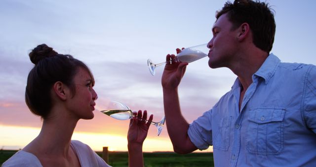A young Caucasian couple enjoys a romantic moment sipping wine at sunset, with copy space. Their silhouettes against the colorful sky create a serene and intimate atmosphere.