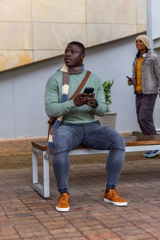 Diverse commuters sitting on bench and walking past while using smartphones in urban plaza