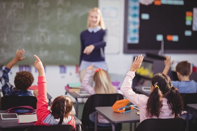 Schoolchildren raising hands in a classroom while a teacher stands at the front. Ideal for educational materials, school websites, and articles about learning and teaching methods. Highlights student engagement and classroom interaction.
