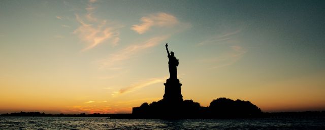 Silhouette of Statue of Liberty stands prominently during sunset with calm waters in the foreground. Ideal for travel blogs, New York tourism promotions, inspirational posters, and educational materials about American landmarks and history.