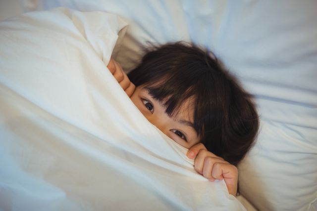 Young boy hiding under a white blanket in bed, peeking out with a playful expression. Ideal for use in articles or advertisements related to children's bedtime routines, family life, home comfort, or playful moments. Perfect for illustrating themes of coziness, innocence, and childhood fun.