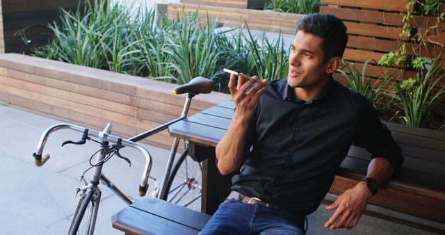 Young man sitting on a bench outside, talking on smartphone, with bicycle near him and green plants in background. Wearing casual outfit, conveying relaxed, modern lifestyle. Perfect for topics related to outdoor activities, urban living, casual communication and eco-friendly transport.