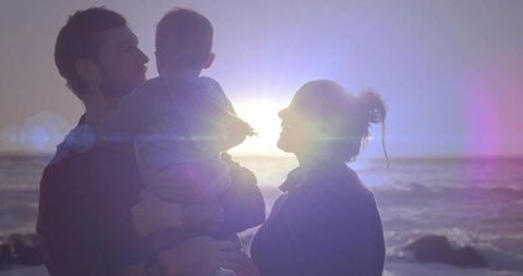 Backlit parents cradling baby at ocean sunset with lens flare and warm silhouette