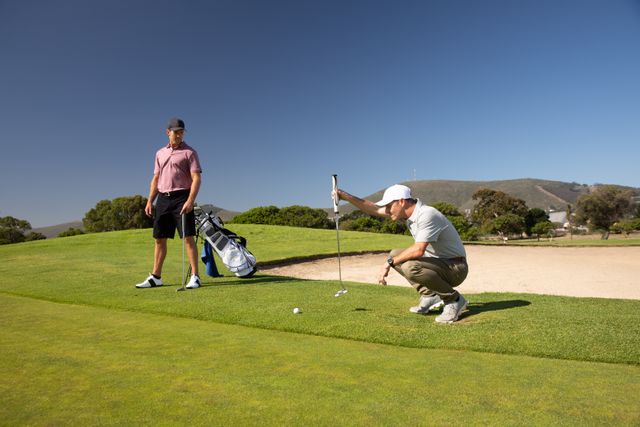 Two male golfers are practicing on a golf course on a sunny day. One man is crouching and checking the position of a golf ball while the other stands nearby. Both are wearing caps and golf clothes. This image can be used for promoting golf courses, sportswear, healthy lifestyle activities, and outdoor hobbies.