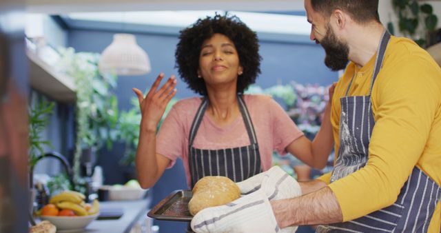 Image of happy diverse couple in aprons baking in kitchen, looking at cooked bread, with copy space. Happiness, communication, inclusivity, free time, togetherness and domestic life.