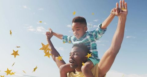 Black father carrying son on shoulders at sunny beach with gold star confetti playful family bonding