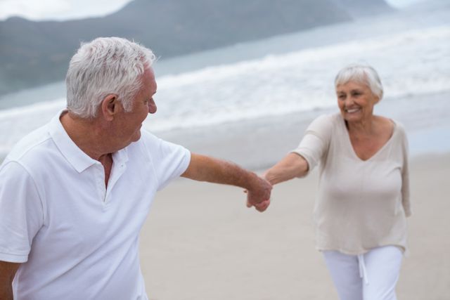 Senior couple holding hands and smiling while walking on the beach. Ideal for use in advertisements, retirement planning materials, travel brochures, and articles about healthy aging and active lifestyles.