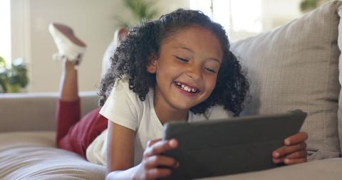 Smiling girl lying on sofa using tablet wearing sneakers in bright cozy living room