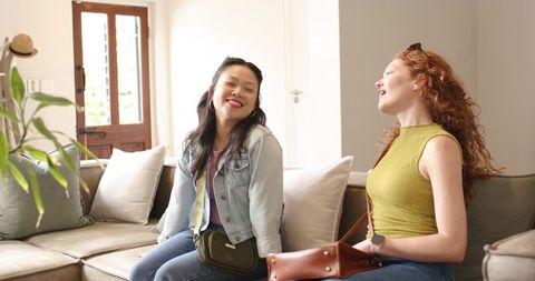 Two diverse female friends laughing on sectional couch in bright modern living room