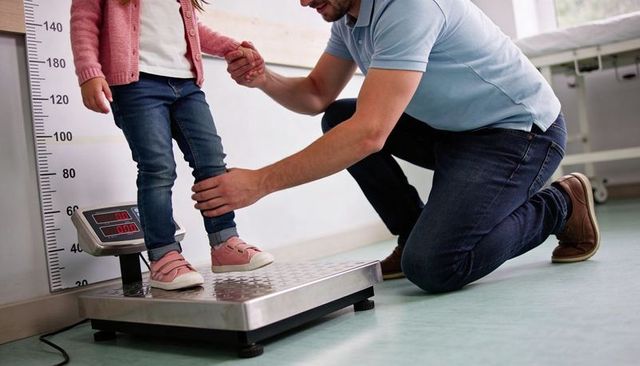 Weighing toddler on clinic scale with height ruler while caregiver steadying during pediatric checku