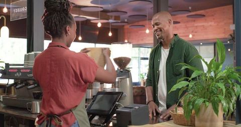 Barista pouring coffee beans into grinder while smiling customer waits at cafe counter