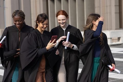 Multicultural women graduates walking together in gowns sharing smartphone and diplomas