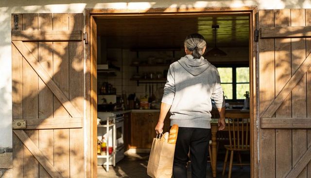 Senior asian man carrying baguettes into sunlit rustic kitchen through wooden barn door