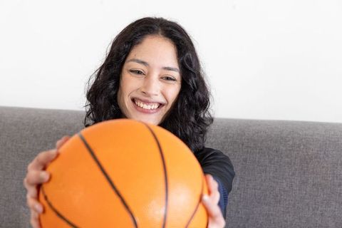 Smiling young woman holding basketball toward camera on grey sofa casual home lifestyle portrait