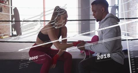 Coach fastening boxer hand wraps on ring apron with analytics overlay in sunlit gym