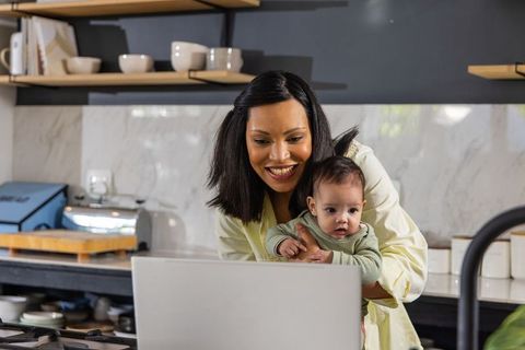 African american mother holding baby while video calling on laptop in modern kitchen