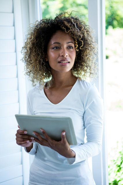 Thoughtful woman standing near the window and using a digital tablet at home