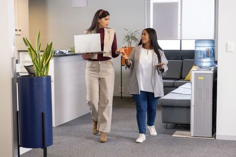 Diverse female coworkers walking and talking in modern office reception carrying laptop