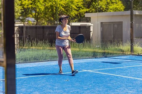 African american woman playing padel on bright blue court wearing visor and activewear